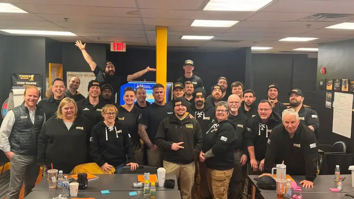 A group of people poses together in a brightly lit office setting, many smiling and wearing matching black shirts with logos. Tables in front hold drinks and office supplies.