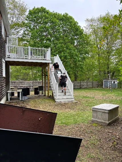Two people are carrying a large item up an outdoor staircase attached to a house, with a grassy yard and trees in the background.
