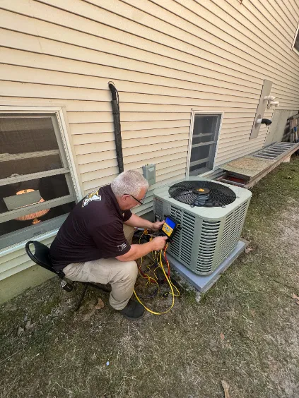 A technician sits on a stool and tests an outdoor air conditioning unit with a digital meter next to a house.
