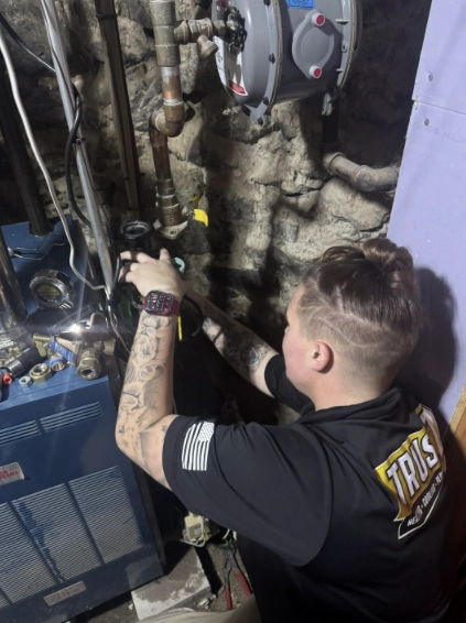 A person with tattoos works on pipes near a blue furnace or boiler in a basement with stone walls, wearing a black "Trust" shirt and a smartwatch.