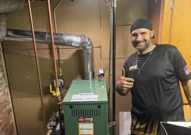 A man in a black shirt stands next to a green boiler in a basement, giving a thumbs-up gesture and smiling at the camera.